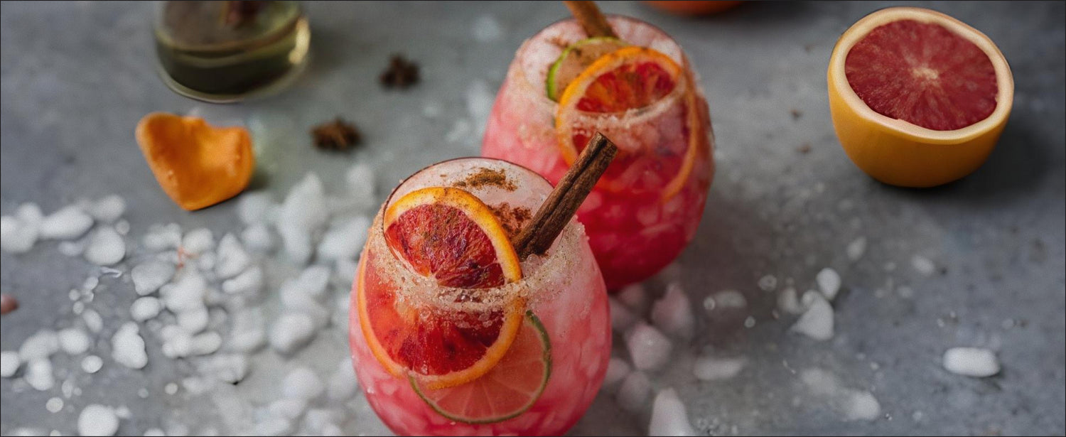 Two pink cocktails with blood orange slices and a cinnamon stick on a gray surface with ice cubes.
